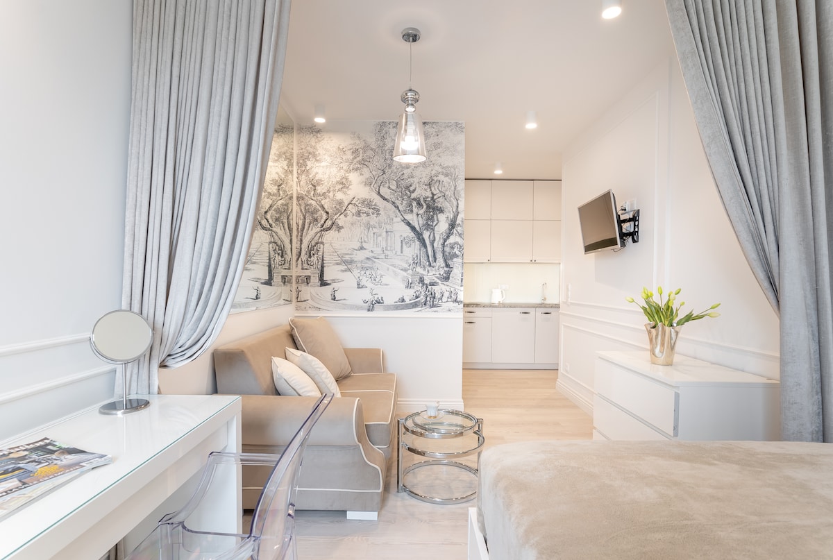 A modern studio space is displayed, featuring a comfortable gray sofa and a sleek glass coffee table. A detailed mural of trees adorns the wall behind the seating area. The kitchenette is visible in the background, complemented by bright lighting and minimalist cabinetry.