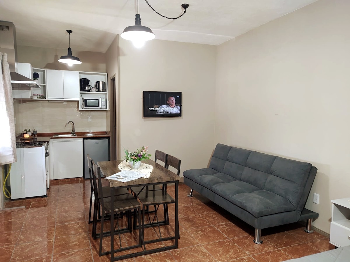 A modern living area is presented with a grey sofa and a dining table accommodating four chairs. The kitchen is visible, featuring essential appliances. Warm lighting is provided by ceiling fixtures, while a television is mounted on the wall, adding to the functional space.
