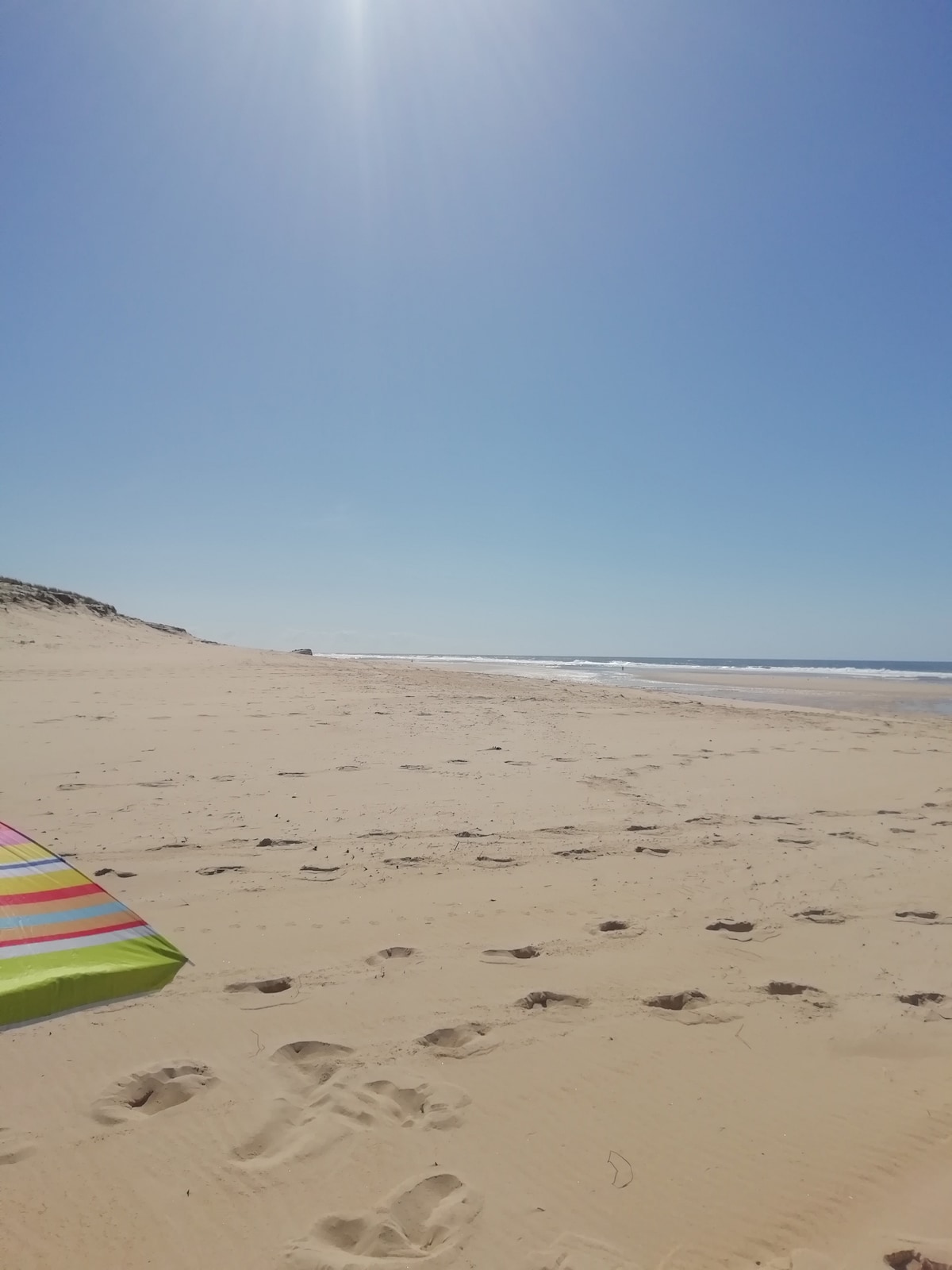 A wide, sandy beach extends under a clear blue sky, leading to gentle waves in the distance. Colorful beach umbrellas are visible in the foreground, providing shade from the sun. The soft sand shows footprints, suggesting a welcoming space for relaxation and enjoyment.