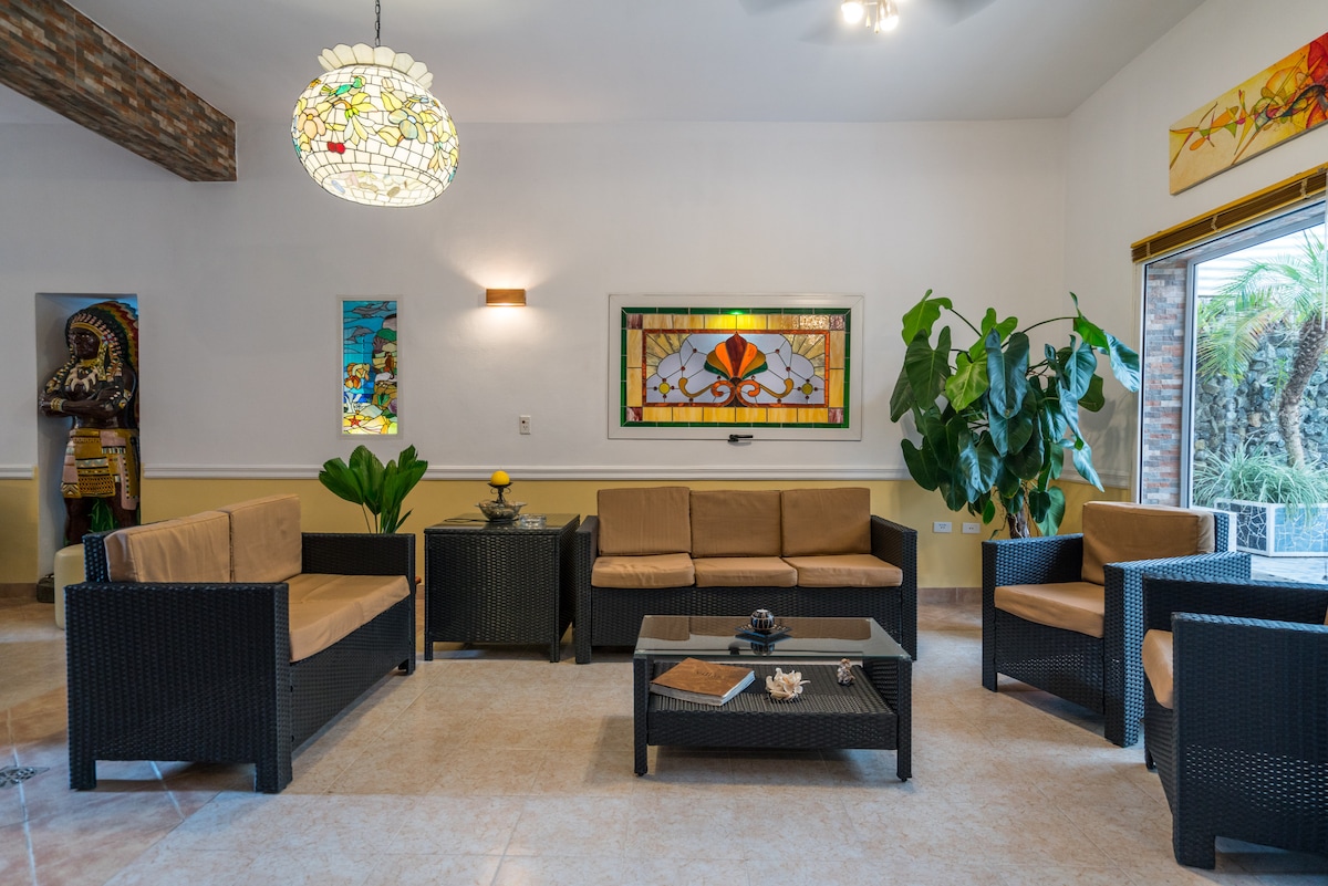 A welcoming lounge area features comfortable wicker seating arranged around a central glass table. A stained glass window and tropical plants add color and vibrancy. Natural light fills the space, enhancing the relaxed atmosphere.