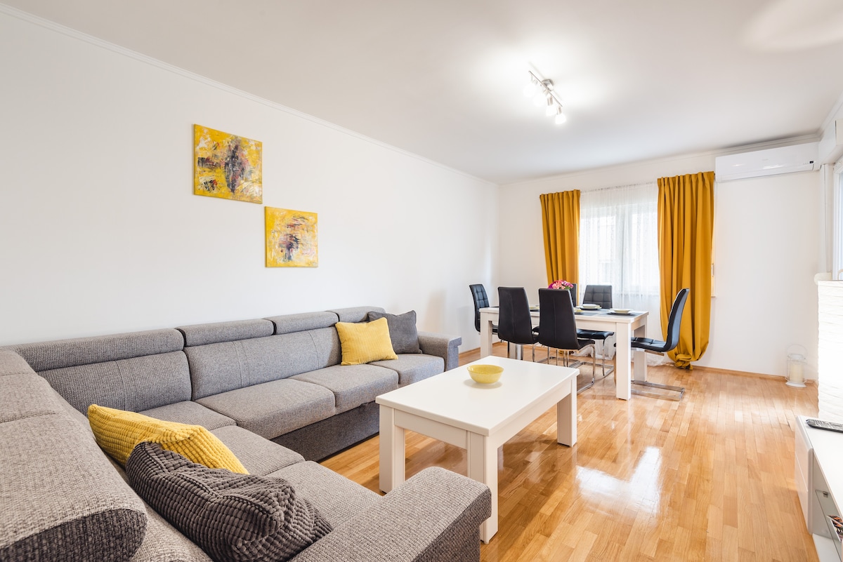 The living room features a spacious gray sofa with yellow accent pillows, complemented by wooden flooring. A white coffee table sits at the center, surrounded by a dining area with a modern table and chairs. Natural light filters through the window, adorned with yellow curtains.