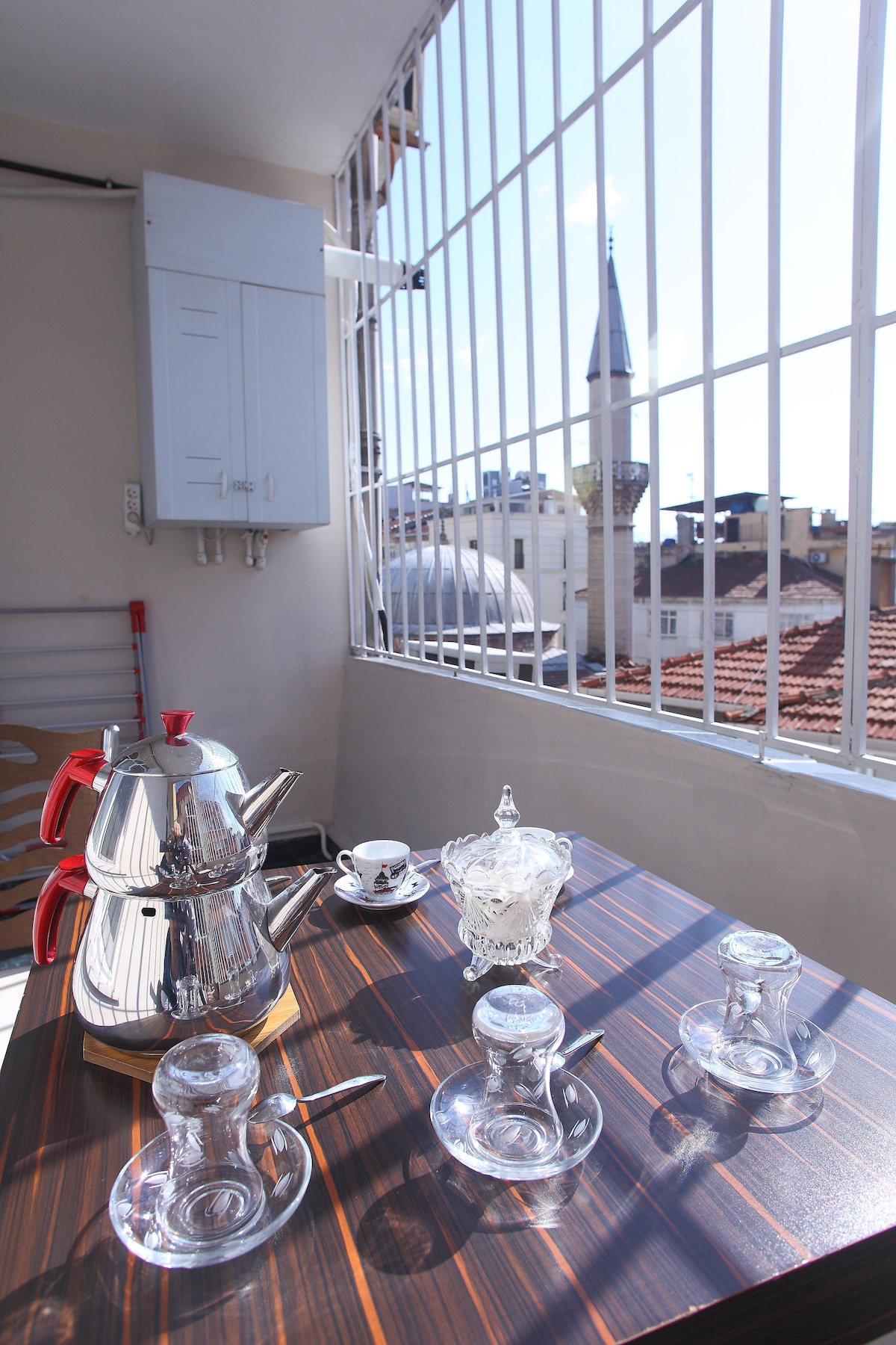 A sunny balcony is depicted, featuring a wooden table set for tea with a traditional Turkish teapot, clear drinking glasses, and a delicate serving dish. In the background, a minaret is visible, framed by the balcony's metal bars.