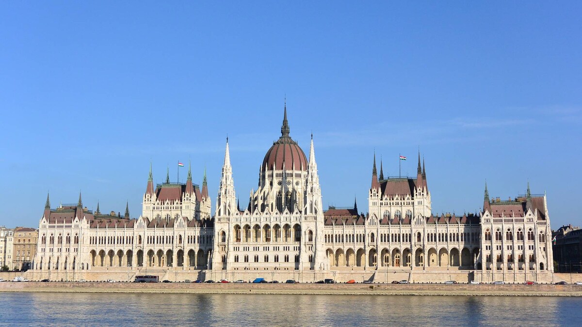 The Hungarian Parliament Building is displayed along the Danube River, showcasing its intricate Gothic Revival architecture. The grand structure features a striking dome, numerous spires, and ornate details, with the river reflecting the building's majestic silhouette under a clear blue sky.