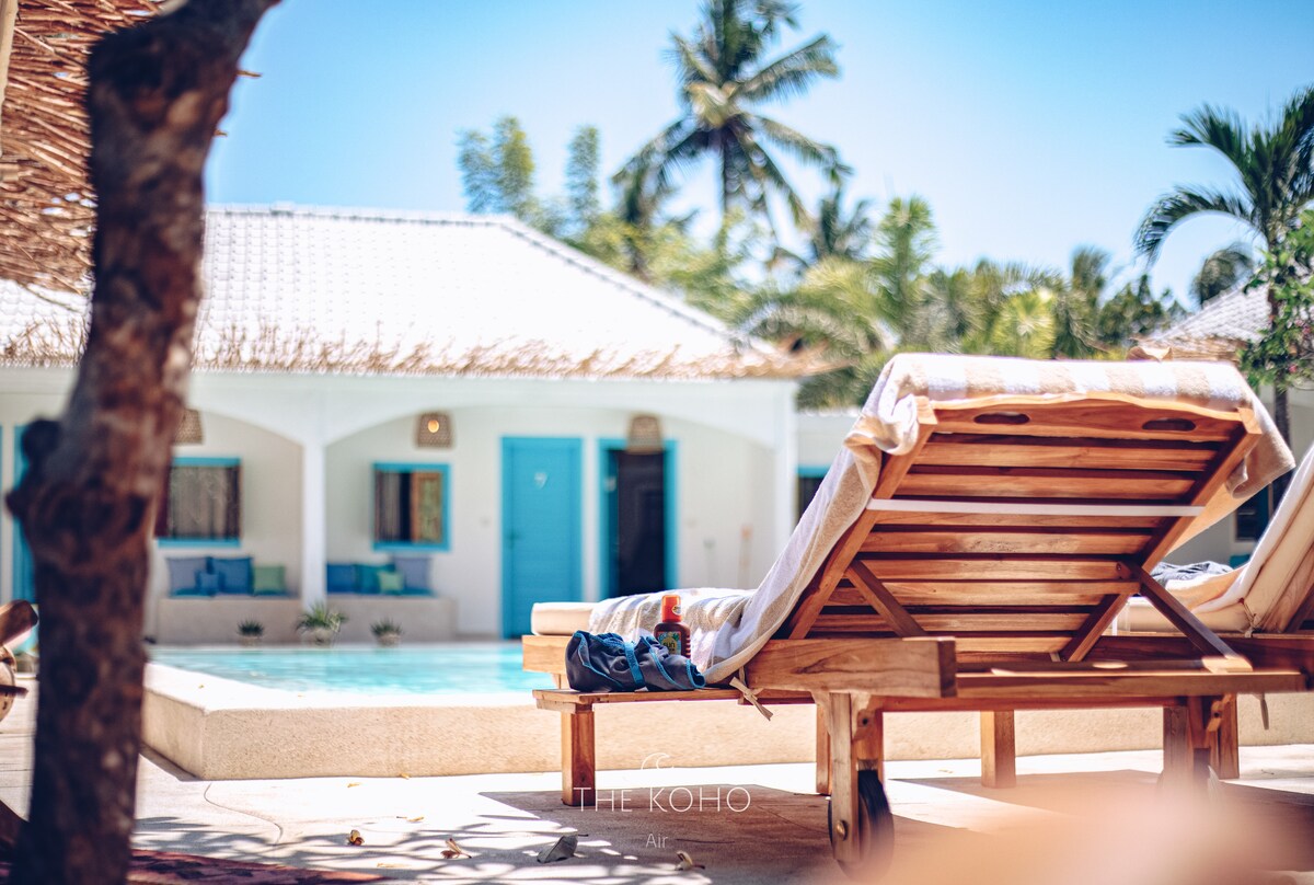 A wooden lounge chair is placed next to a swimming pool, complementing the vibrant outdoor setting. Lush green palm trees provide a tropical backdrop, while a quiet white building with blue accents is visible in the distance, contributing to a relaxed atmosphere.
