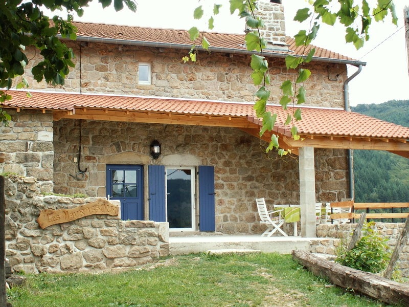 A traditional stone house with a terracotta roof is shown, featuring blue shutters framing the entrance. A porch area is visible, furnished with a small white table and chairs, overlooking lush greenery and distant hills.