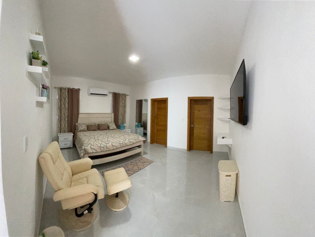 A spacious bedroom features a large bed with neutral bedding, accompanied by a cozy recliner and a small side table. A wall-mounted television is positioned across from the bed. The light-colored walls and floors contribute to a bright and open atmosphere.