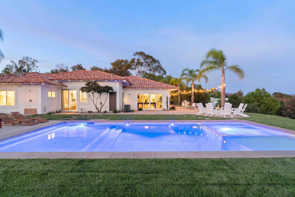 A modern Mediterranean home with a spacious outdoor area features a shimmering pool surrounded by lush grass. Comfortable lounge chairs are placed nearby, and twinkling string lights are seen illuminating the patio area, enhancing the relaxing ambiance. Palm trees line the property, adding a tropical touch.