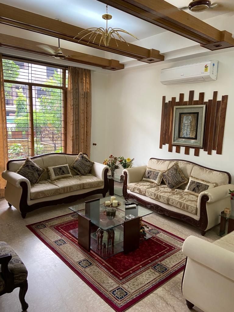 A comfortable living area features two elegant sofas adorned with decorative cushions and a glass coffee table at the center. A plush area rug adds warmth, while natural light filters through a large window lined with sheer curtains. Decorative accents enhance the inviting atmosphere.