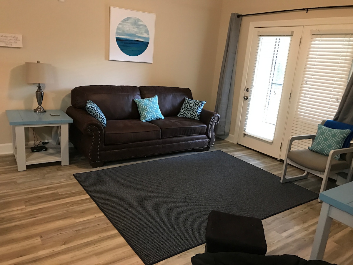 A comfortable living area is furnished with a brown sofa and two decorative cushions in blue. A light blue table sits beside the sofa, and a second seating option in gray with blue accents is positioned nearby. Natural light enters through the sliding glass doors, enhancing the cozy atmosphere.