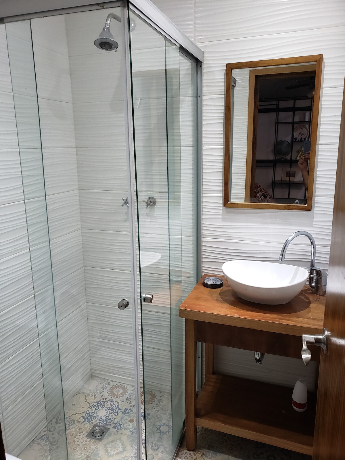 The bathroom features a glass-enclosed shower with a modern showerhead and a small wooden vanity. A round basin sits atop the vanity, complemented by a framed mirror. The tiled floor exhibits decorative patterns, adding character to the space.