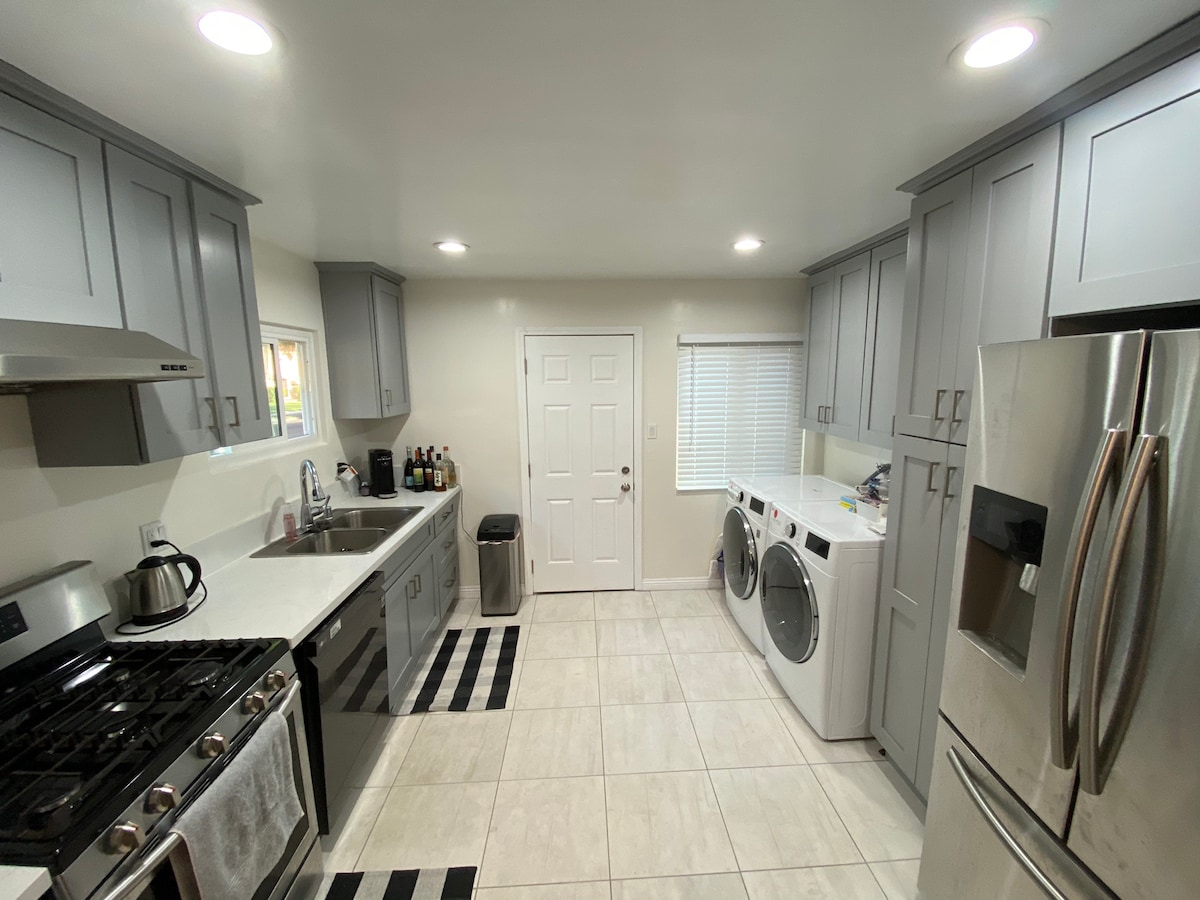 The kitchen features gray cabinetry with modern stainless steel appliances, including a gas stove and refrigerator. A washing machine and dryer are located near the back door, providing convenience for guests. Bright overhead lighting illuminates the space, creating a functional cooking environment.