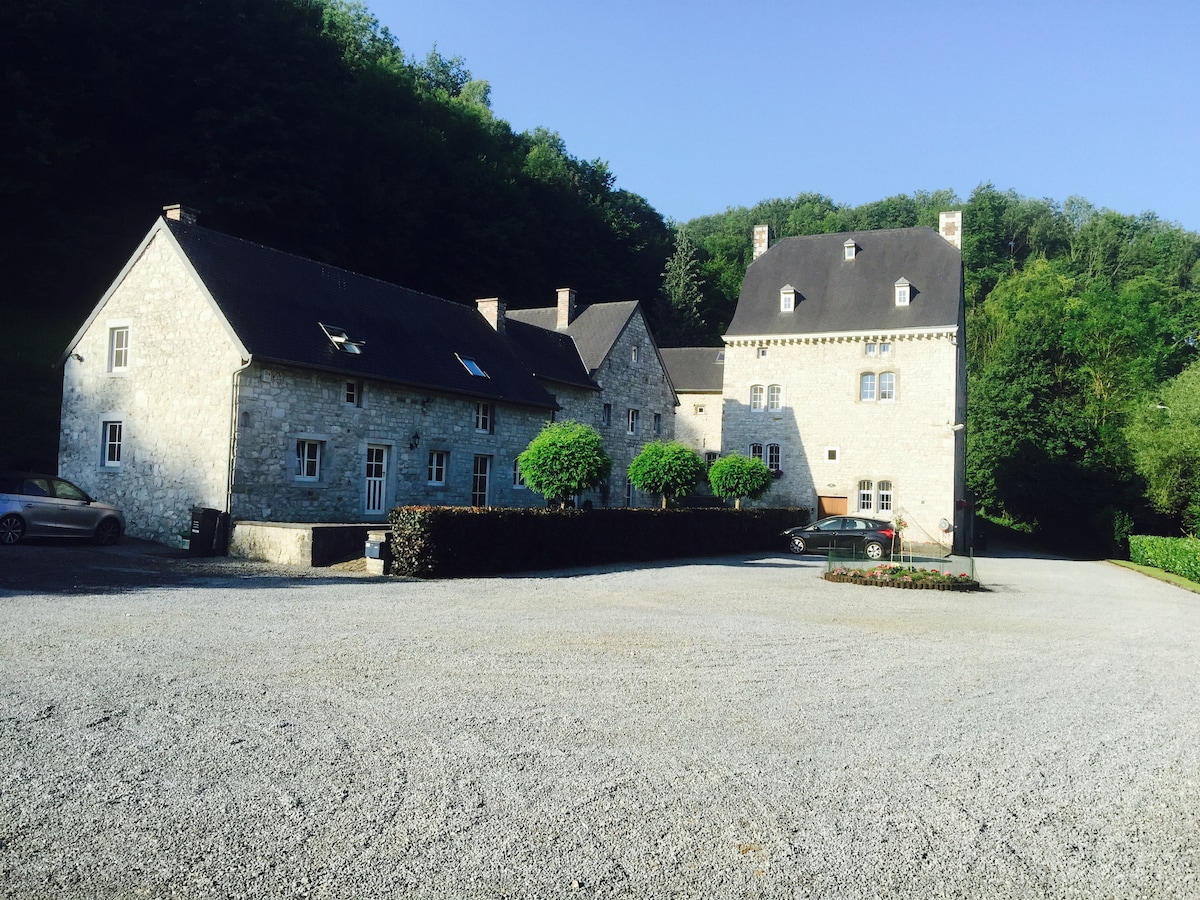 A large stone house from the 18th century is set against a lush green hillside. The building is comprised of multiple sections, featuring a gravel parking area with several parked cars. Neat hedges and small trees surround the entrance, enhancing the inviting exterior.