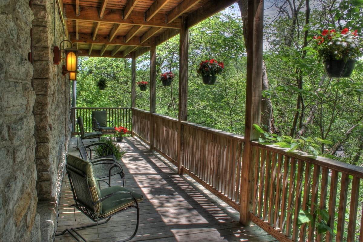 A covered terrace features wooden flooring and a railing, offering views of surrounding greenery. Two comfortable chairs are positioned along the space, with hanging planters filled with vibrant flowers providing seasonal color.