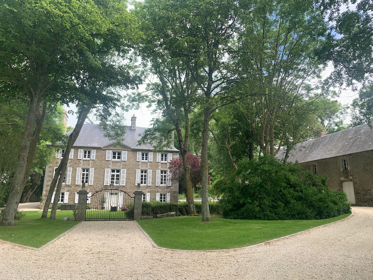 A charming stone property is surrounded by lush greenery, featuring multiple windows and a welcoming entrance gate. A driveway leads through the manicured lawn, providing a sense of serenity. Tall trees offer shade and complement the natural setting.