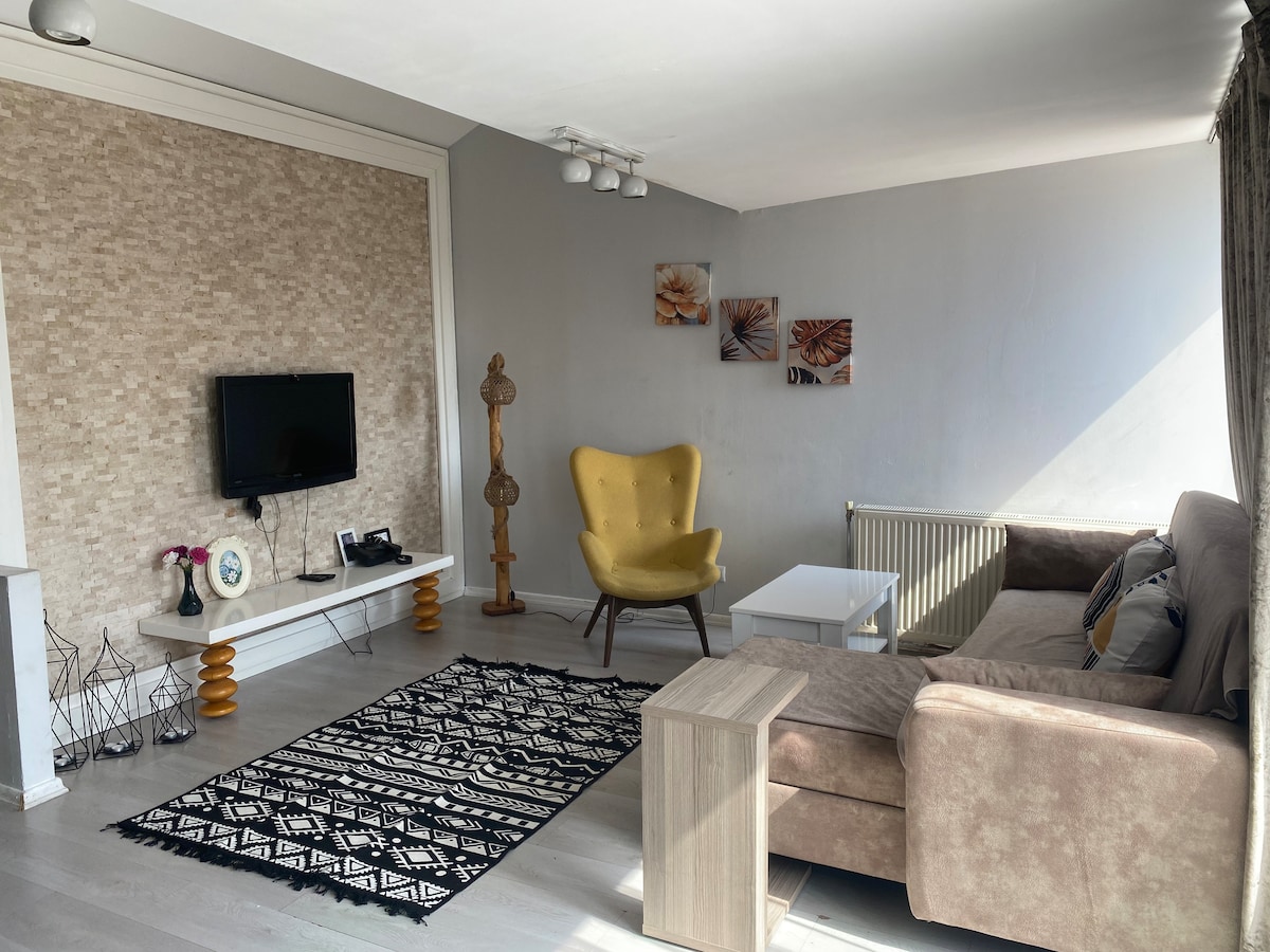 A bright living area features a large window allowing natural light to fill the space. A comfortable sofa and a decorative yellow chair are positioned around a small coffee table. An area rug adds texture to the floor, and artwork decorates the wall above.