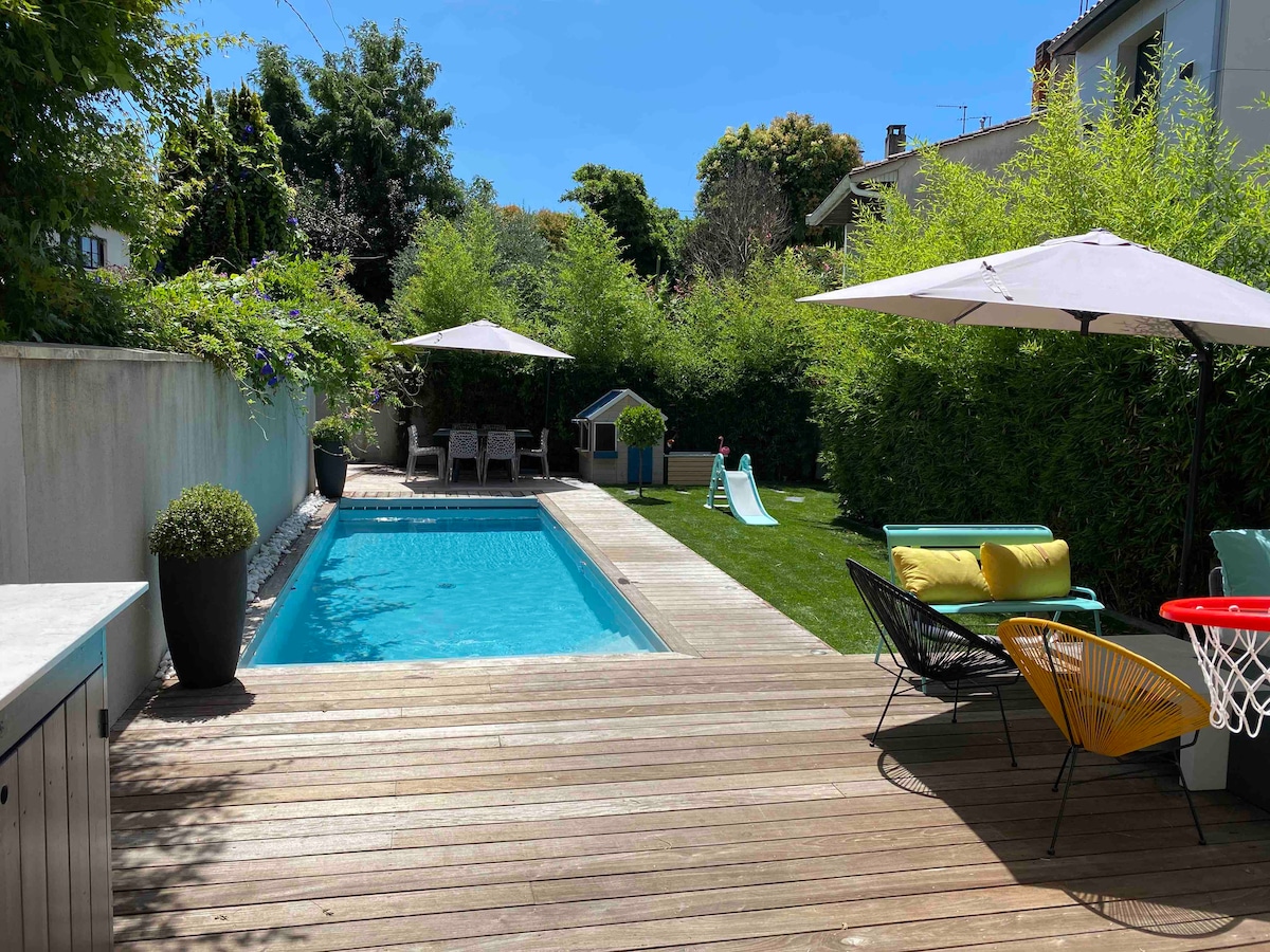 A private outdoor space is showcased with a sparkling pool bordered by a wooden deck. Comfortable seating areas with umbrellas offer shade, while a grassy area features a slide and a basketball hoop. Lush greenery surrounds the setting, enhancing the tranquil atmosphere.