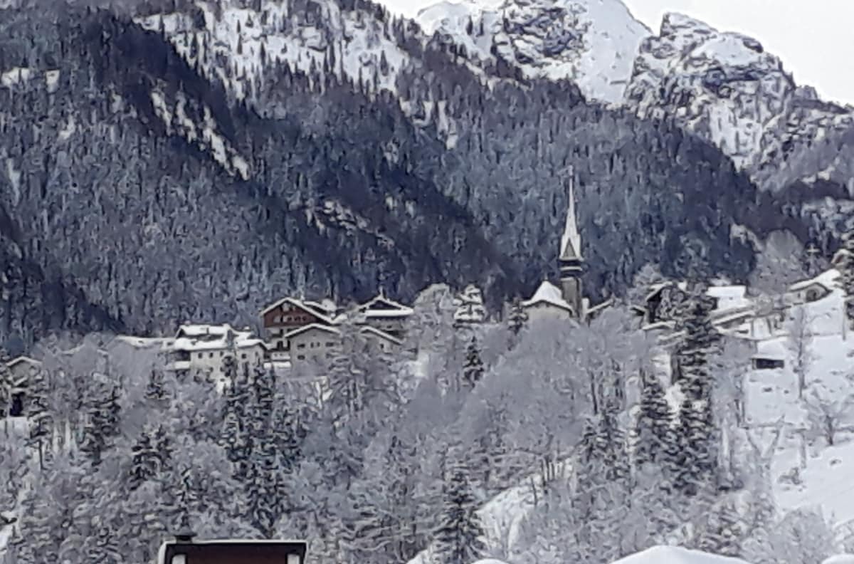 A tranquil winter landscape is depicted, featuring snow-capped mountains in the background and a quaint village nestled among frosted evergreen trees. A church steeple rises above the buildings, which display rustic architecture harmonizing with the surrounding alpine scenery.