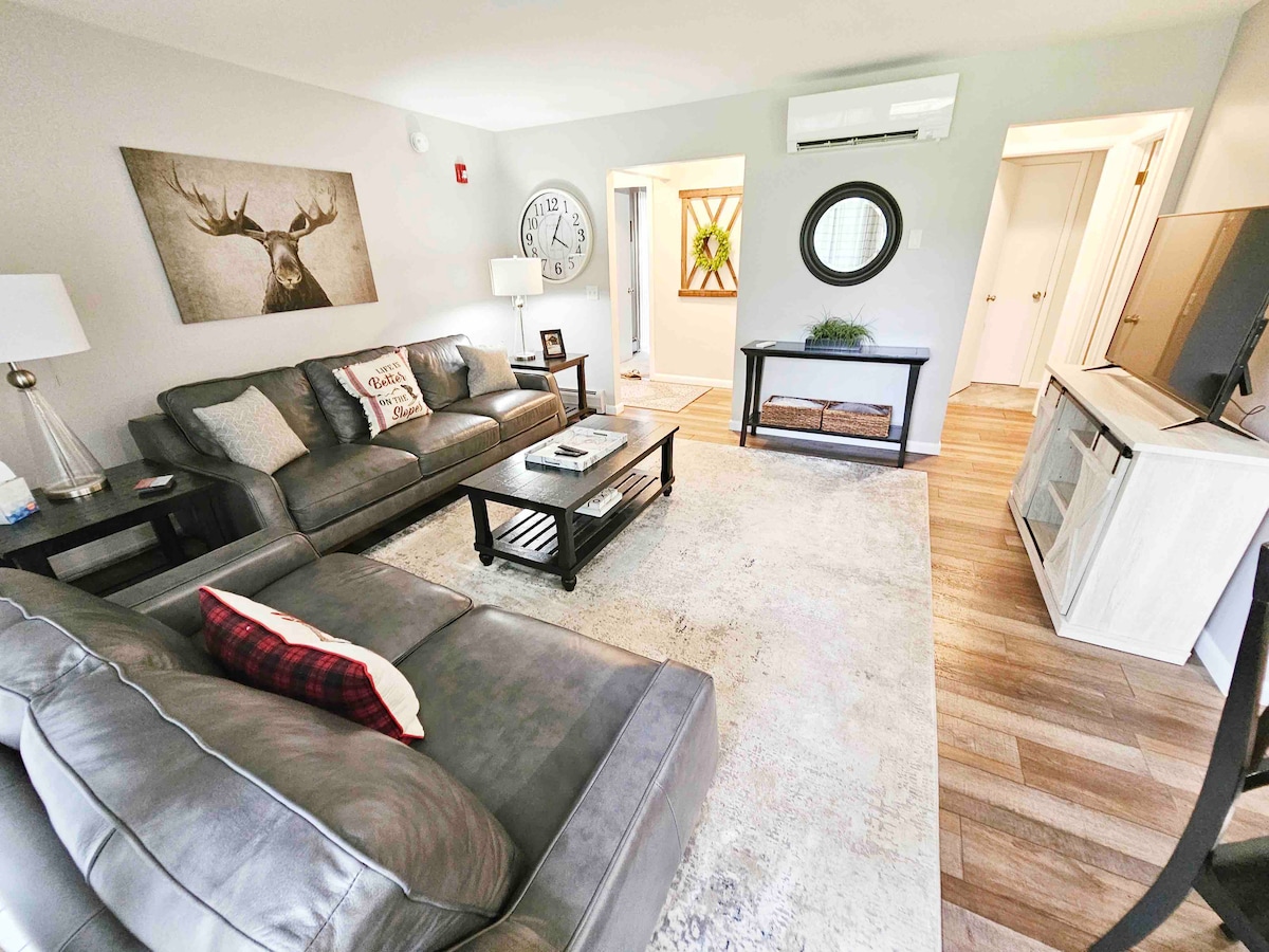 A welcoming living area is designed with comfortable seating, including a large gray sofa and armchairs. A round wall clock and an artwork featuring a deer add character. A small table holds decorative items, while natural light enters through the nearby entrance.