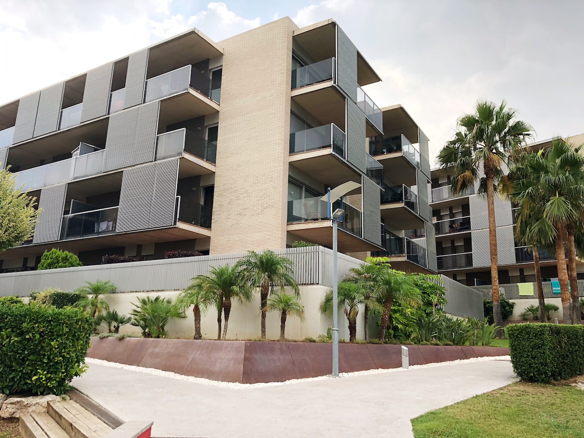 The apartment complex features modern architecture with multiple balconies. Lush palm trees and landscaped greenery create a welcoming ambiance in the foreground. A wide pathway leads to the entrance, highlighting the clean lines and contemporary design of the building.