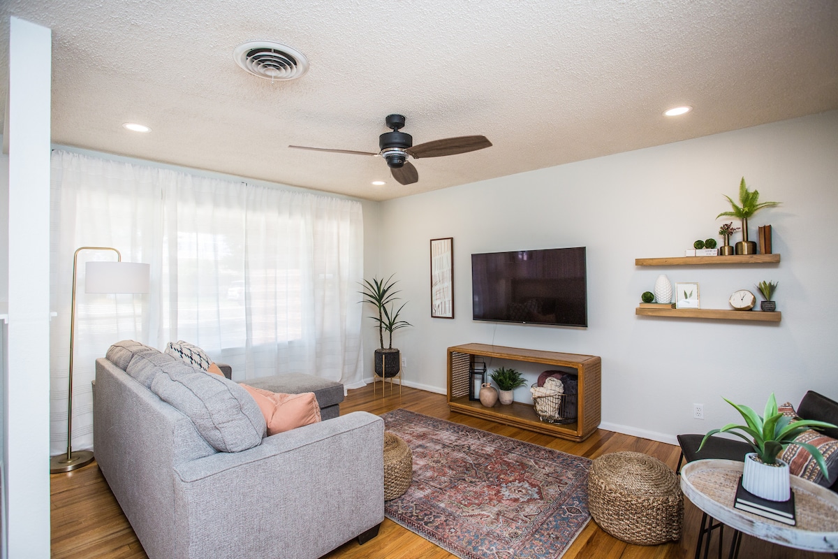 A living room is showcased with a light-colored sofa complemented by throw pillows. A television is mounted on the wall above a low media console adorned with decorative items. Natural light filters through sheer curtains, and indoor plants add a touch of greenery.
