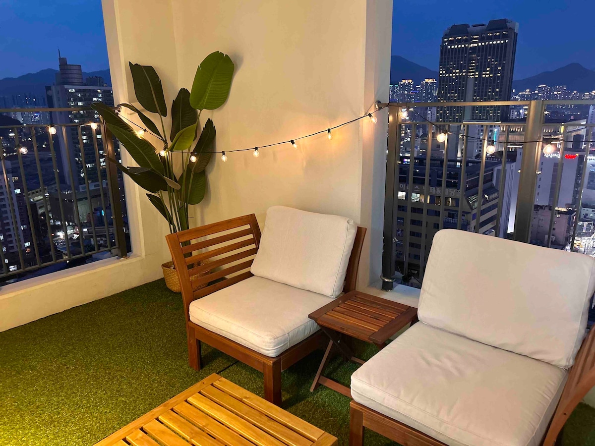 A cozy terrace is visible, featuring two wooden chairs with soft cushions and a small side table. A potted plant adds a touch of greenery, while string lights softly illuminate the area against a backdrop of city lights after dusk.