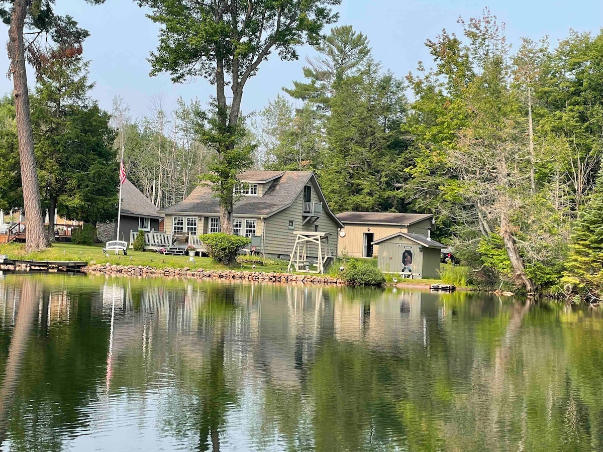 A tranquil lakeside view features a family-friendly cabin surrounded by trees. The structure is reflected in the calm water, highlighting the serene environment. A gazebo and various outdoor seating areas are visible along the shore, enhancing the natural landscape.