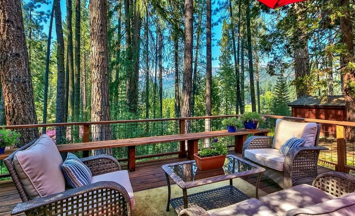 A spacious deck is framed by tall trees, providing a scenic view of the surrounding nature. Comfortable seating is arranged around a glass table, complemented by potted plants. An umbrella offers shade, enhancing the inviting atmosphere for relaxation and enjoyment of the outdoors.