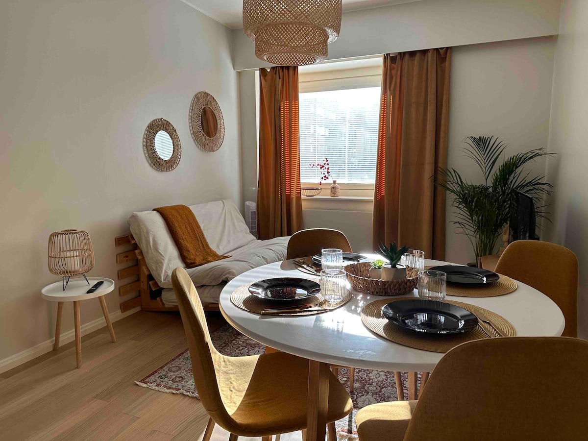 A dining area is shown with a round table set for four, featuring dark plates and glassware. A futon sofa is positioned against the wall, accompanied by woven decor and potted greenery. Natural light filters through curtains, enhancing the warm tones of the room.