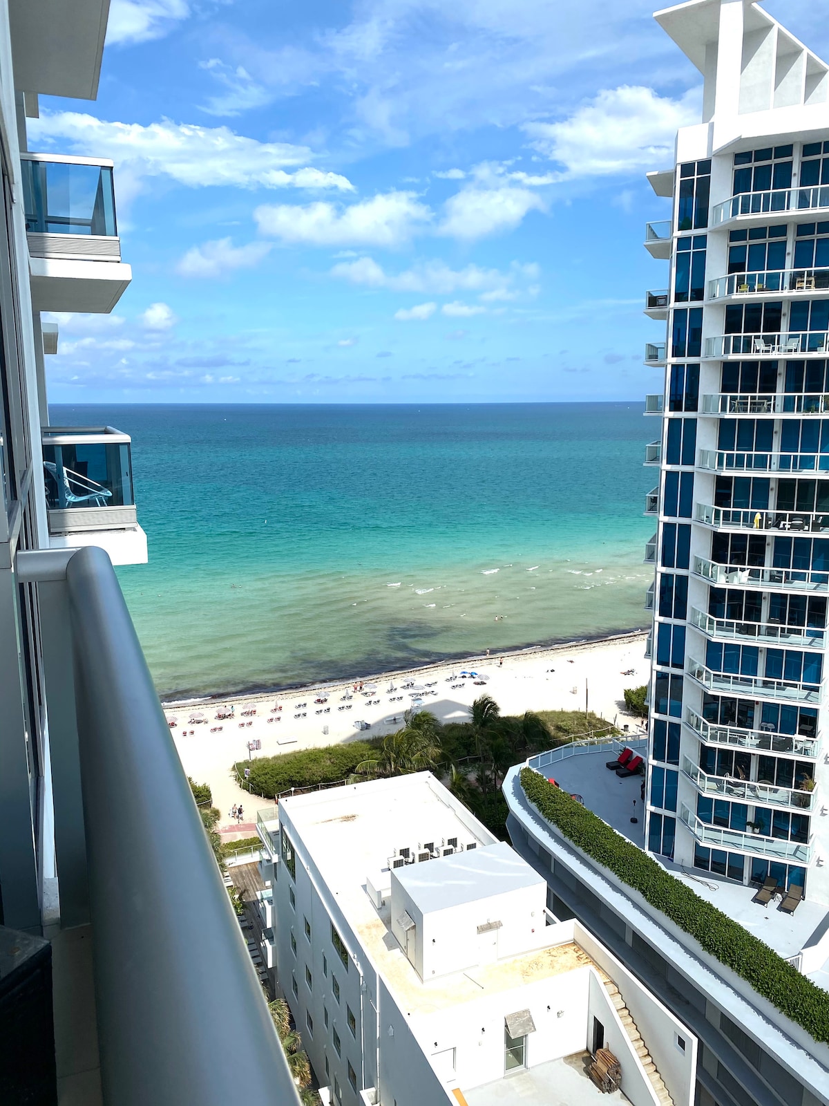 An expansive view showcases the ocean with varying shades of blue, framed by modern high-rise buildings. A white sandy beach with arranged loungers and umbrellas is visible below, while clear skies enhance the bright and inviting atmosphere.