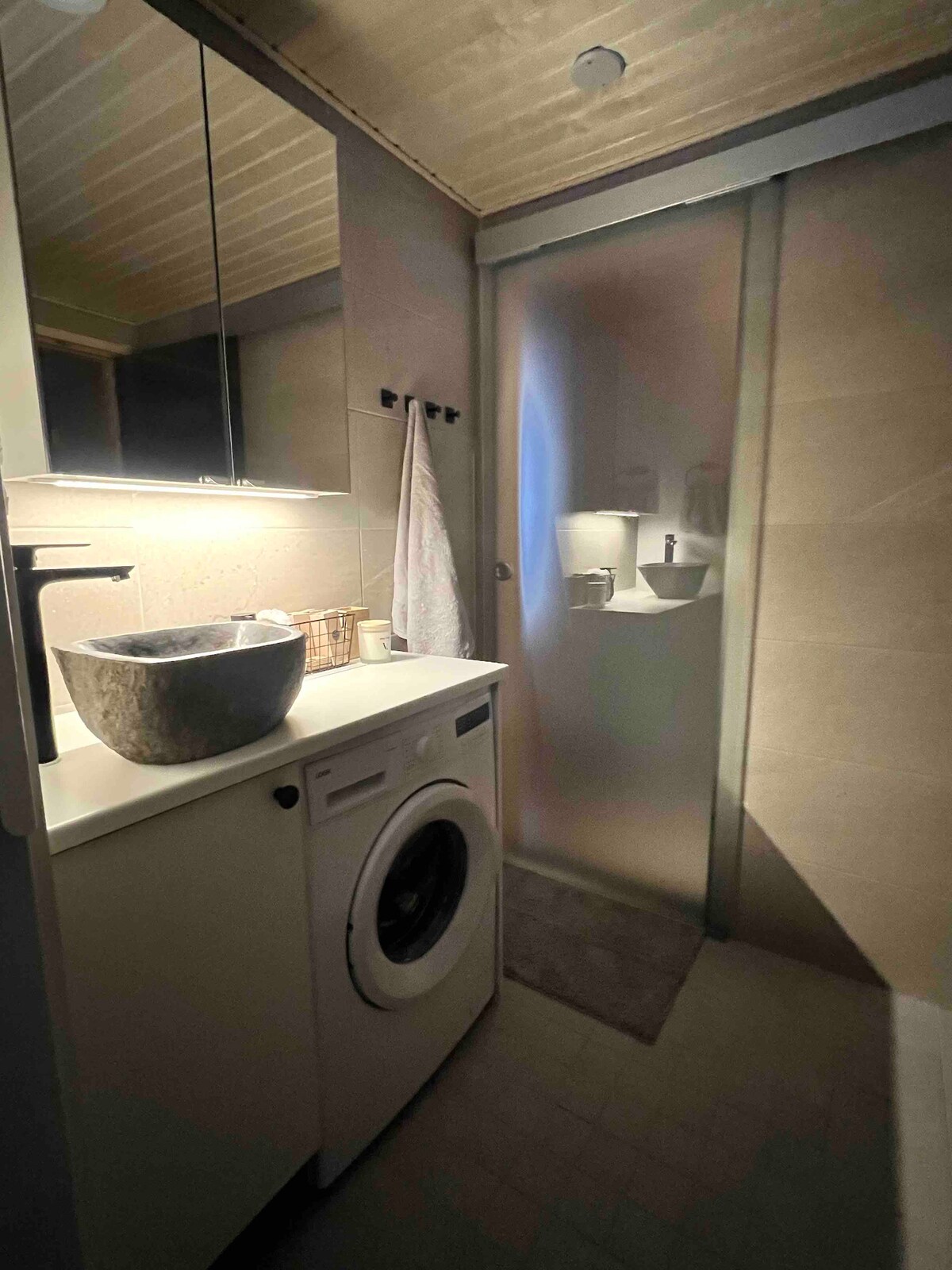 A modern bathroom is equipped with a washing machine and a large basin. A frosted glass panel separates the shower area, while soft lighting highlights the neutral-toned tiles. A towel is hung on a hook, and a small basket sits on the countertop.