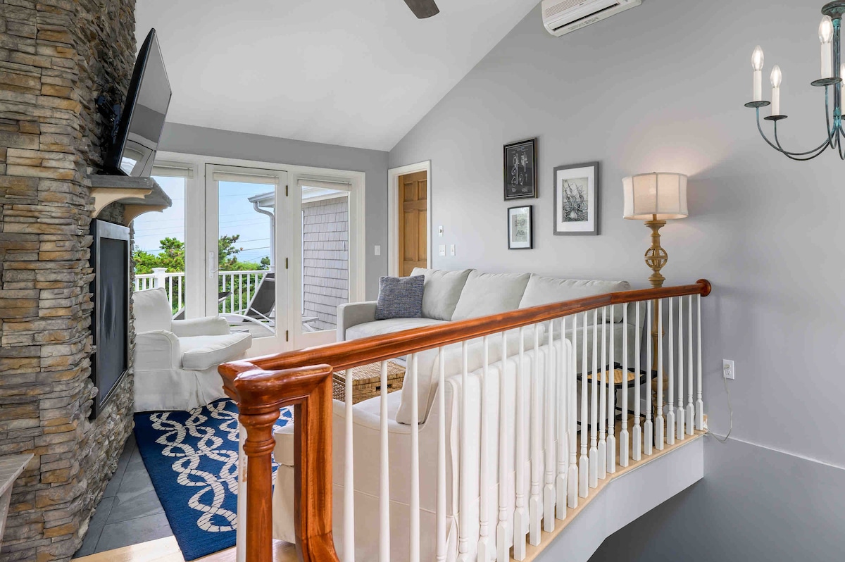 A spacious living area features a sleek stone fireplace and a large flat-screen TV mounted on the wall. Natural light floods in through glass doors leading to a deck, while a neutral-colored sofa is complemented by a decorative area rug.