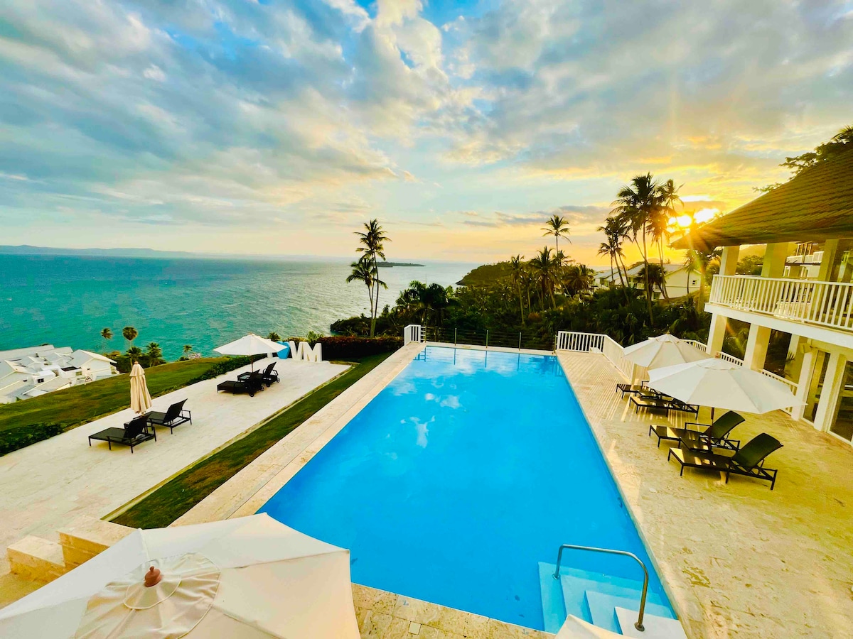 A vast pool area is visible, surrounded by sun loungers and umbrellas. The tranquil water reflects the sky's hues as the sun sets over the ocean. Lush greenery and palm trees frame the scene, enhancing the serene coastal atmosphere.