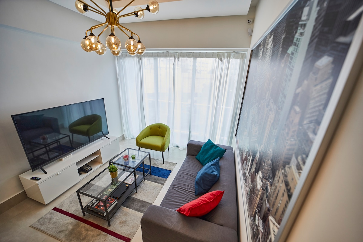 A living area features a large television mounted on a white entertainment unit. A gray sofa adorned with colorful cushions is positioned beside a glass coffee table. A bright green chair provides additional seating. Soft natural light enters through large windows with sheer curtains.