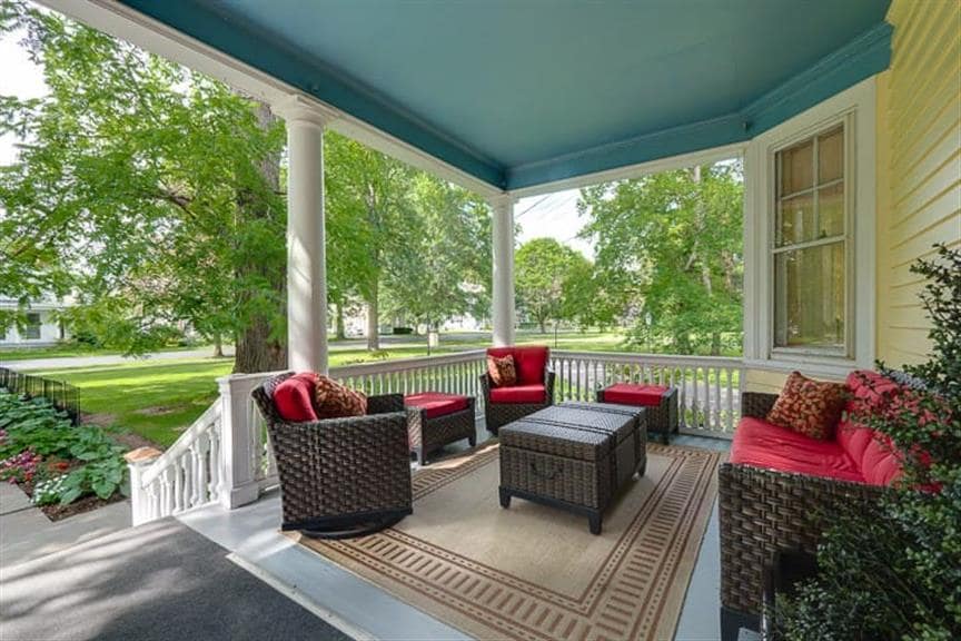 A spacious porch area features a seating arrangement of four dark wicker chairs, each adorned with red cushions, around a small, rectangular table. Lush green trees and flower beds are visible, contributing to a serene outdoor setting.