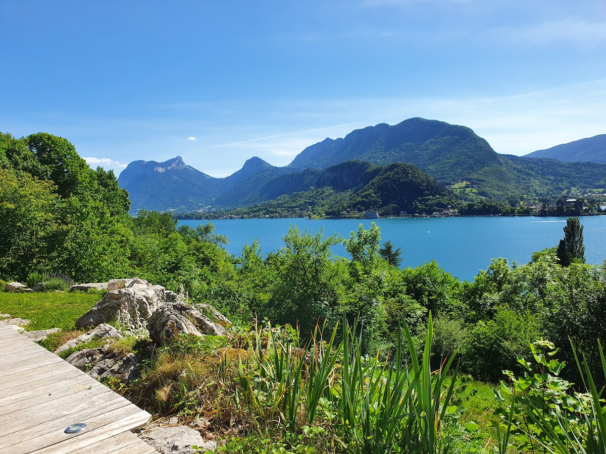 A panoramic view of Lake Annecy is visible, surrounded by lush greenery and distant mountains. The sunlit scene showcases a wooden deck along with natural rock formations and vibrant foliage in the foreground.