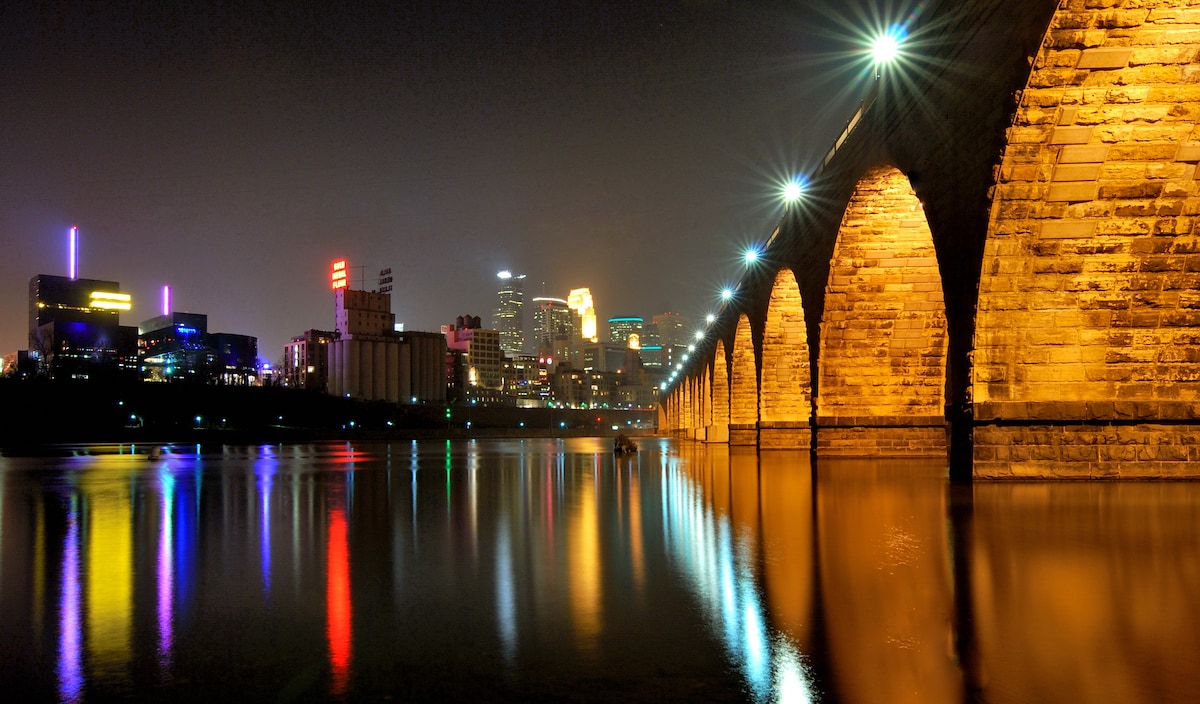 The image showcases the Stone Arch Bridge, illuminated by warm lights reflecting off the calm waters below. The Minneapolis skyline serves as the backdrop, with colorful lights on nearby buildings adding a dynamic touch to the night scene.