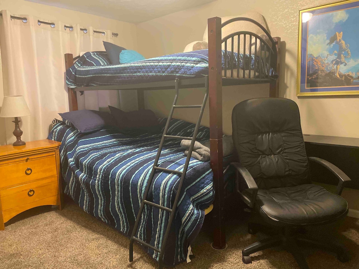 A cozy private room features a full bed with a bunk above it, layered with a blue-striped blanket. A ladder provides access to the top bunk. Nearby, a black rolling chair and a wooden nightstand are positioned against a neutral wall, contributing to a functional layout.
