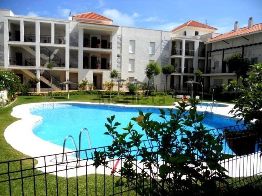 A spacious outdoor area features a large swimming pool surrounded by manicured lawns and small trees. The building showcases a modern design with balconies overlooking the pool, creating a serene environment. Poolside seating and umbrellas are visible for relaxation.