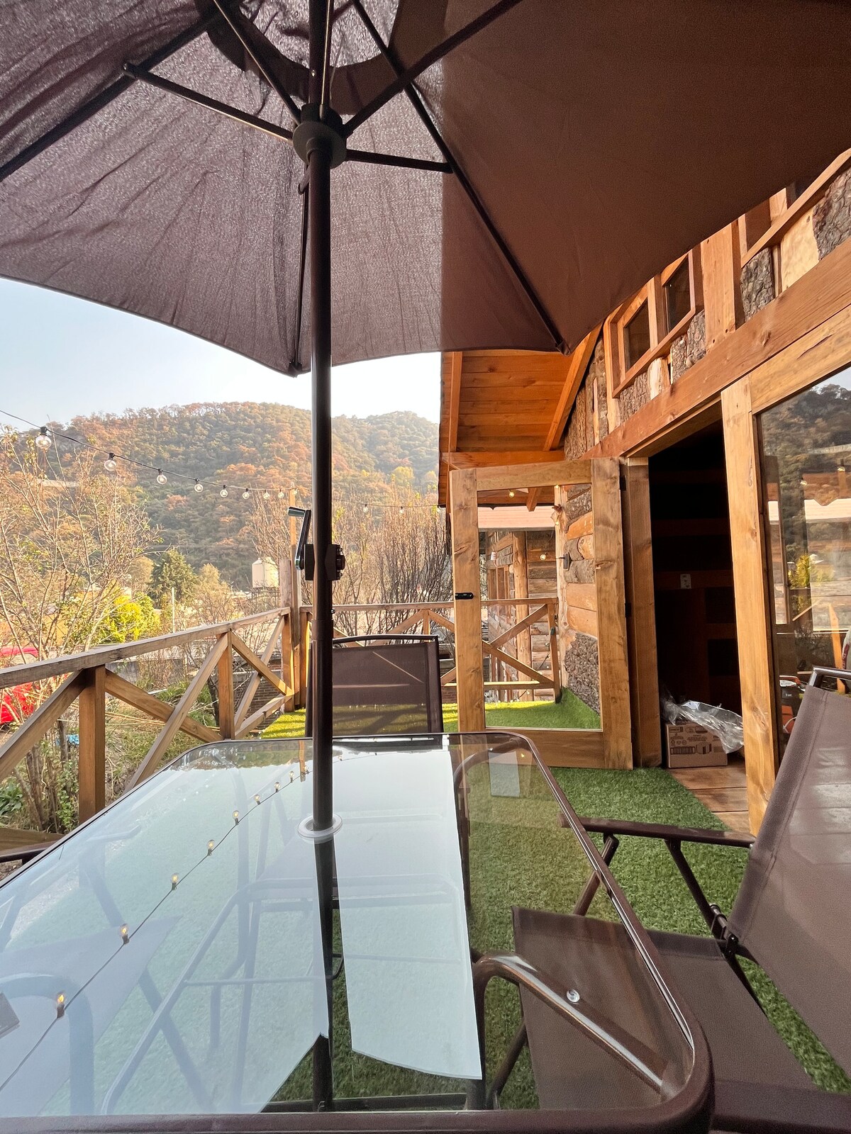 An outdoor dining area is shown, featuring a glass table with a large umbrella. Two shaded lounge chairs are positioned nearby, overlooking the scenic mountainous landscape. Wooden structures and warm tones are present in the background, enhancing the tranquil atmosphere.