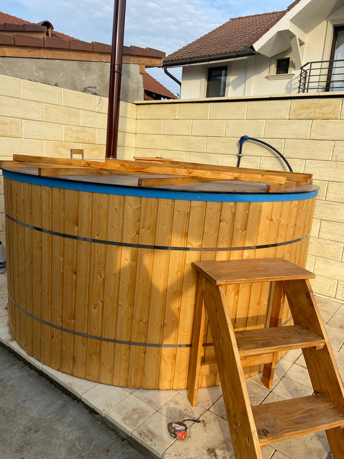 A rustic wooden hot tub is displayed, featuring a round shape and a blue trim. A wooden ladder is positioned beside it, allowing easy access. The surrounding area shows stone walls and tiled flooring, contributing to an inviting outdoor space.