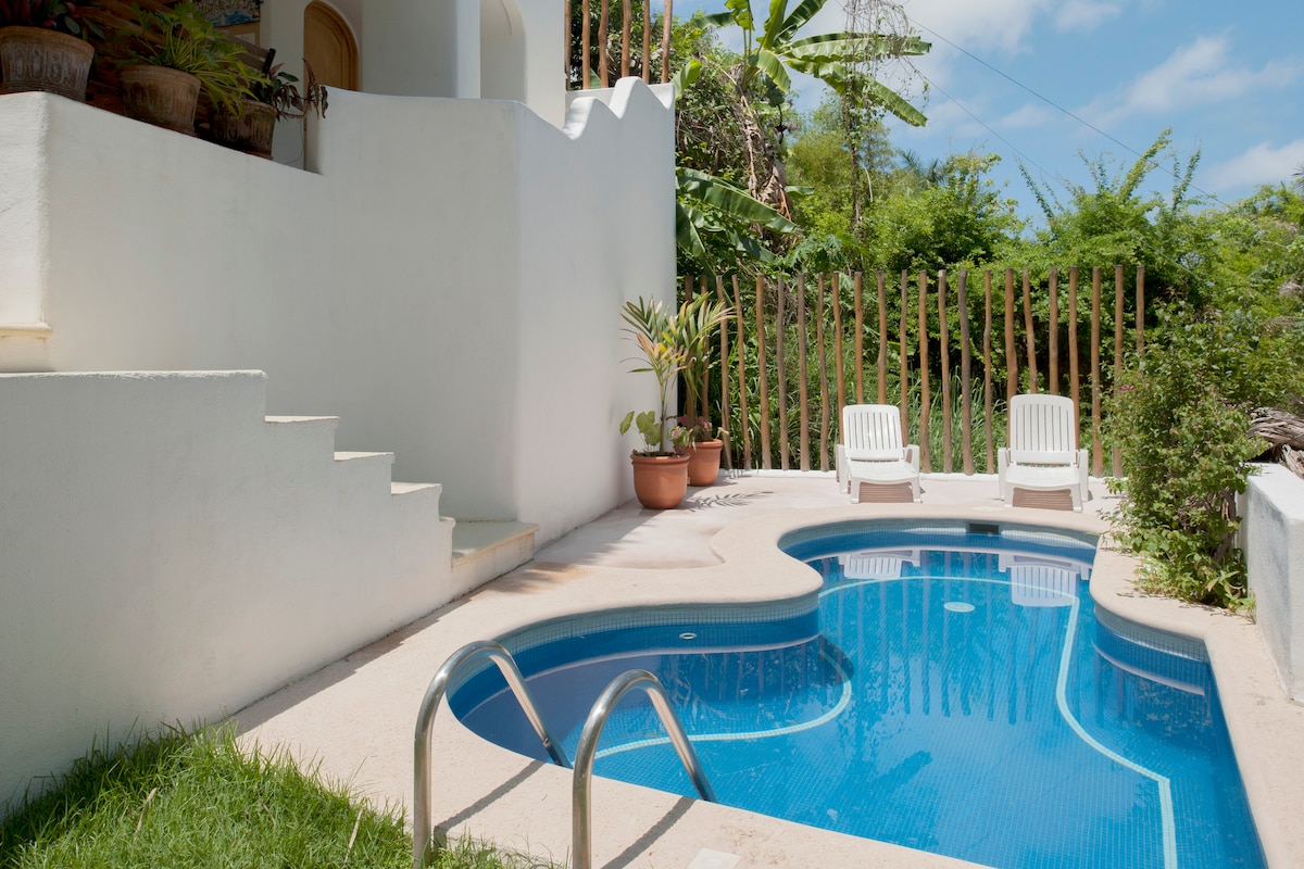 A shaped swimming pool is featured in a lush outdoor setting. Two white lounge chairs are positioned beside the pool, and a small staircase leads up to additional greenery. Walls of textured white surround the area, enhancing the tropical ambiance.