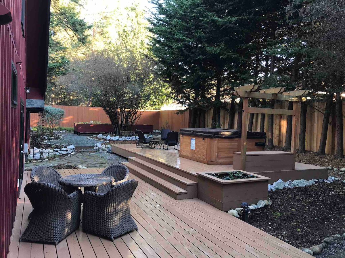 An outdoor deck is presented with a hot tub and seating area, surrounded by lush greenery and wooden fencing. A fire pit is visible on the side, along with pathways lined by stones, enhancing the tranquil atmosphere for relaxation and gathering.