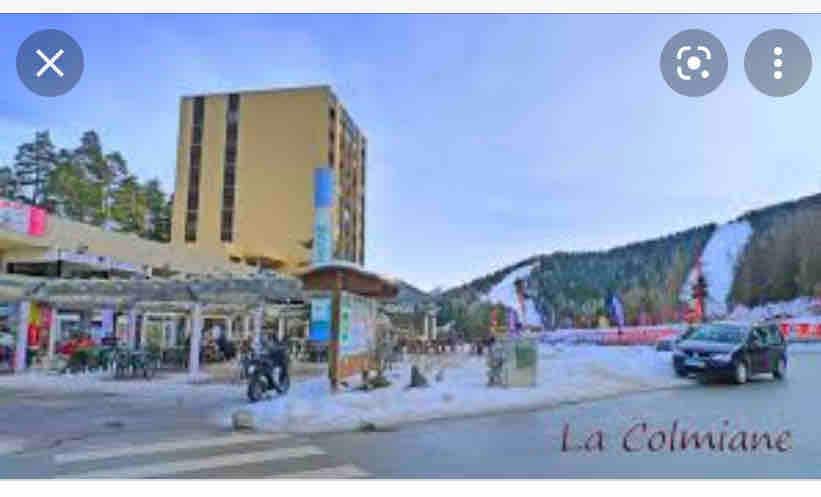 A view of the ski station La Colmiane features a building surrounded by snow-covered terrain. In the foreground, a parking area and a mix of bicycles and vehicles are visible. The backdrop showcases the ski slopes, with trees lining the edges.