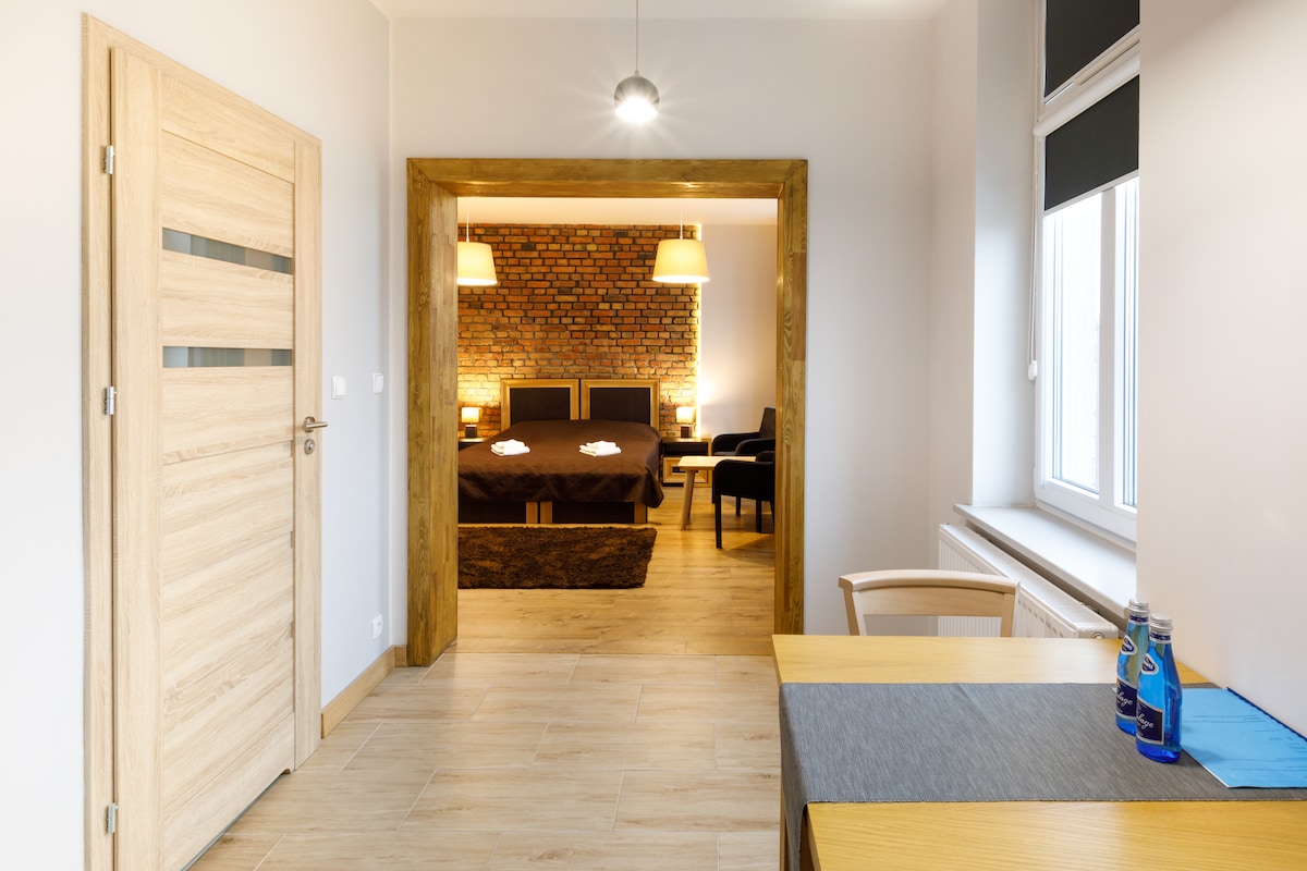 An entrance area opens into a combined living and sleeping space, featuring a wooden-framed doorway. Two spacious beds are visible in the background, complemented by a textured brick wall. A small dining table is positioned in the foreground, along with bottled water.