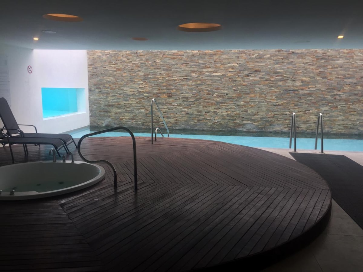 An indoor pool area features a textured stone wall and large windows, allowing natural light to illuminate the space. Relaxing lounge chairs are positioned on a wooden deck beside a circular hot tub, creating a calm environment for guests to unwind.