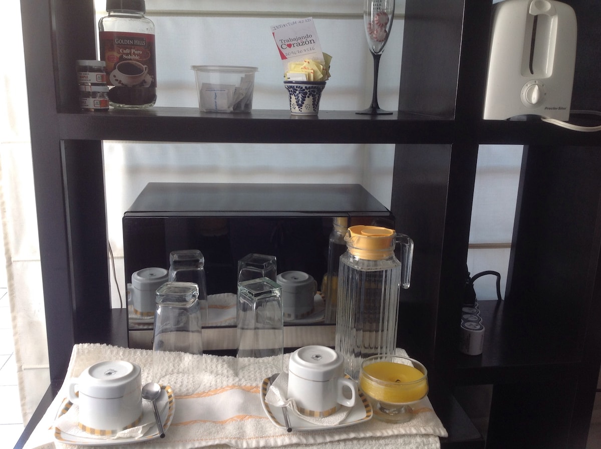 A black shelving unit displays an array of items, including a microwave, toasters, and glassware. Two cups on trays are set alongside a glass jar containing yellow liquid. A small decorative bowl and a bottle are placed on the bottom shelf, with additional kitchenware visible.