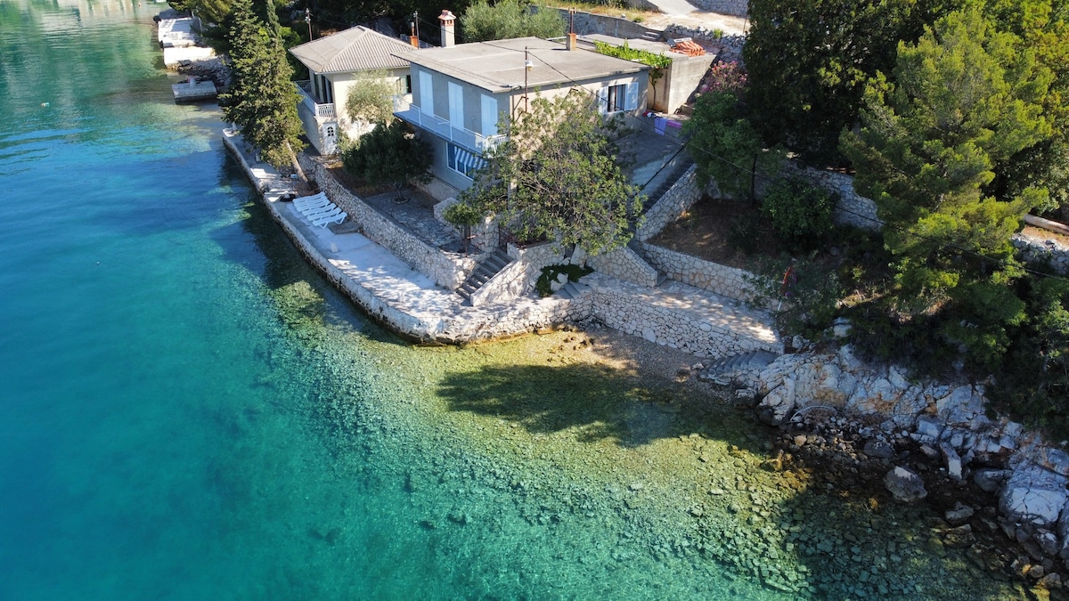 A seaside house is positioned near clear turquoise waters, with stony paths leading to the water's edge. Lush greenery surrounds the property, while the sun casts gentle shadows across the rocky shore, creating a serene environment for relaxation.