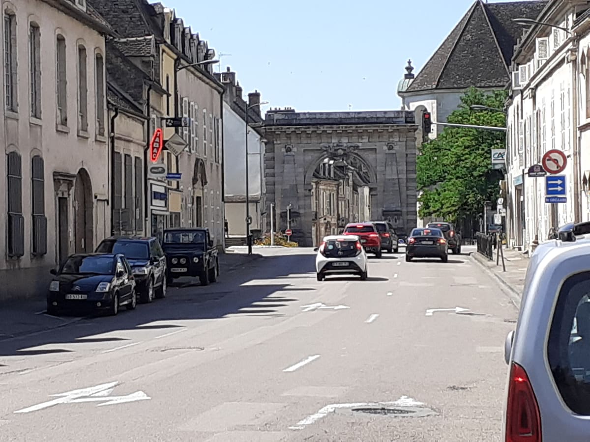 A wide street lined with historic buildings features parked cars on either side. In the distance, a prominent archway is visible, leading to a brick pathway. Traffic signals and street signs are present, indicating the urban setting of the area.