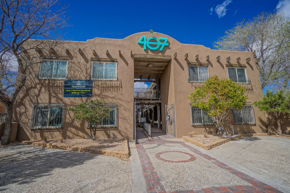 The exterior of a two-story adobe-style building is presented, featuring a welcoming entrance with the number 407 prominently displayed. Surrounding greenery complements the structure, and large windows provide a glimpse into the interior. A patterned stone pathway leads to the door, set against a clear blue sky.