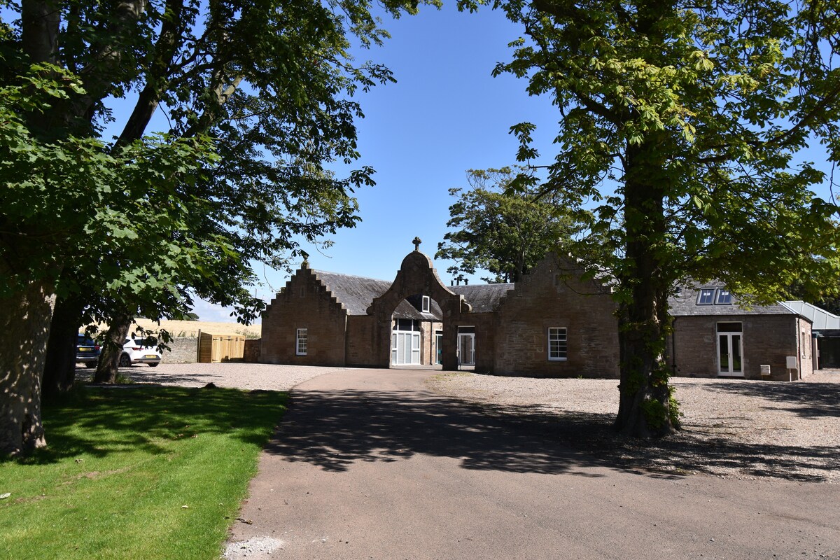 The exterior of the Tack Room @ Panbride House is showcased, featuring a stone stable block with a distinctive gabled roof and central entrance. Lush trees flank the property, offering shaded areas, while a gravel driveway leads up to the building. The spacious grounds allow for ample outdoor space.